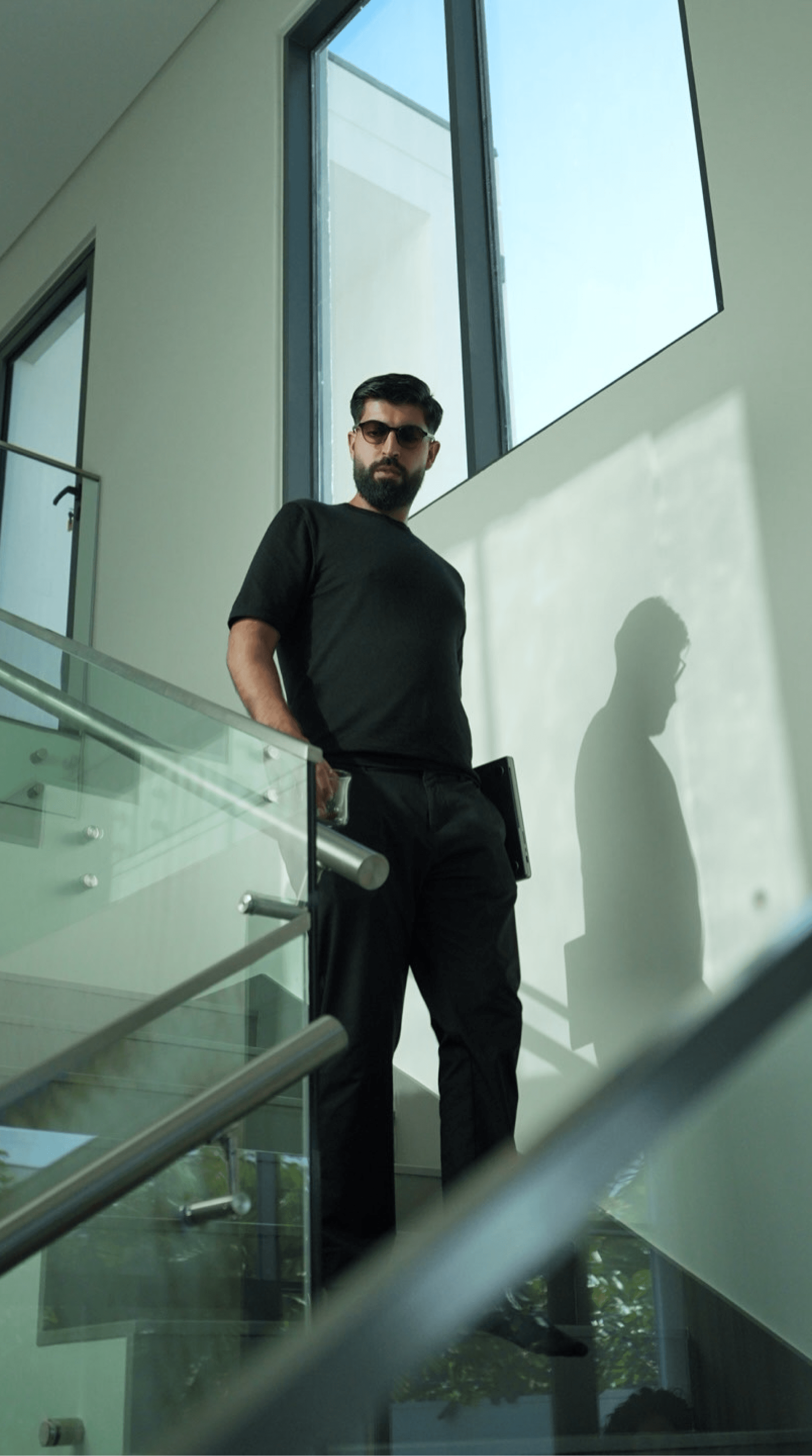 Faruk Ilkhan standing on a staircase landing in a bright modern house, wearing black, with sunlight falling across the wall behind him